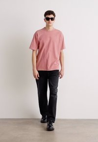 Lyserød bomulds t-shirt, korte ærmer, afslappet pasform, parret med sorte denim jeans. Modellen bærer sorte solbriller og sorte sko.