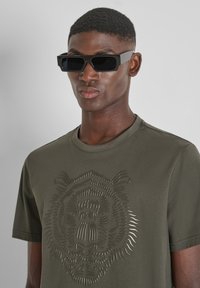 Olijfgroen t-shirt met een textuurontwerp van een leeuwengezicht; korte mouwen. Zwarte rechthoekige zonnebril met donkere lenzen.