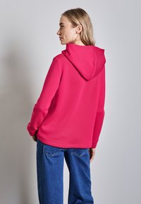 Sweatshirt à capuche fuchsia avec une texture lisse, doté d'une coupe décontractée, de manches longues et d'une poche kangourou. Associé à un jean bleu.
