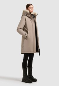 Parka impermeabile color beige chiaro con cappuccio foderato in peluche, tasche laterali e chiusura con zip. Abbinato a leggings neri e stivali robusti con lacci.