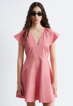 Žena s dlhými kučeravými vlasmi, ktorá má na sebe čierne slnečné okuliare a ružové krátke šaty s hlbokým výstrihom do V na jednoduchom bielom pozadí.