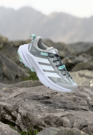 Zapatilla de correr gris con rayas blancas, cuerpo de malla texturizada, acentos turquesa y suela resistente, flotando sobre una superficie rocosa.