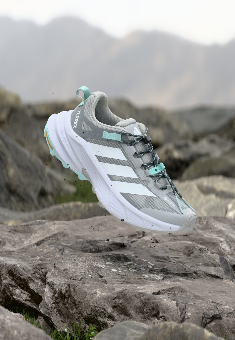 Scarpa da corsa grigia con strisce bianche, corpo in mesh testurizzato, accenti turchesi e suola robusta, che fluttua sopra una superficie rocciosa.