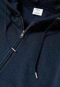 Sweat à capuche zippé bleu marine en tissu doux, doté d'une fermeture éclair argentée, de cordons de serrage avec embouts métalliques, et d'une étiquette à l'intérieur du col avec les détails de la marque.