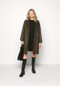 Cappotto in pelle verde oliva con colletto in pelliccia marrone, due bottoni, abbinato a un vestito nero e stivali neri al ginocchio. Borsa nera in mano.