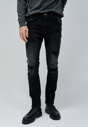 Jeans de denim negros con parches desgastados y un ajuste ceñido, combinados con botas negras de cordones. El material tiene una textura suave.