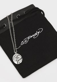 Srebrna ogrlica s visećim digresijskim privjeskom s kružnim oblikom in zapletenim dizajnom zmaja, položena na črn oblaček tkanine z belim napisom.