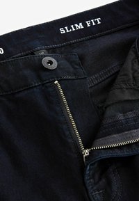 Donkere denim slim-fit jeans met een ritssluiting, knoopsluiting en een zachte textuur. Witte "SLIM FIT" tekst op de tailleband, geen patronen.