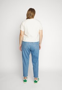 Personne aux cheveux châtain clair portant une chemise blanche à manches courtes, un jean bleu et des baskets blanches et vertes, debout de dos.