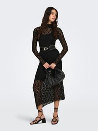 Vestido de encaje negro con mangas largas, cuerpo ajustado y dobladillo asimétrico. Se lleva con un cinturón con tachuelas, un bolso negro y sandalias negras con tiras.