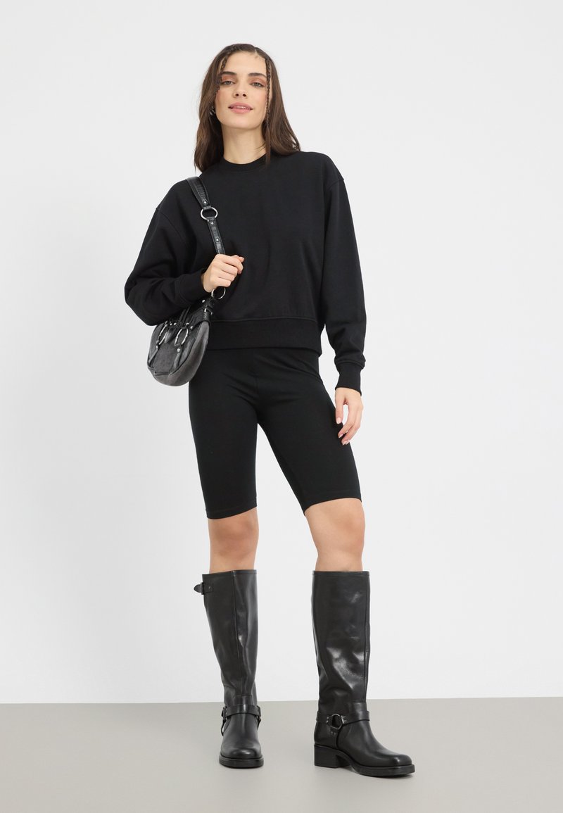 Sweatshirt nero a maniche lunghe e corto, pantaloni biker neri e stivali alti in pelle nera. Modella che tiene una piccola borsa nera.