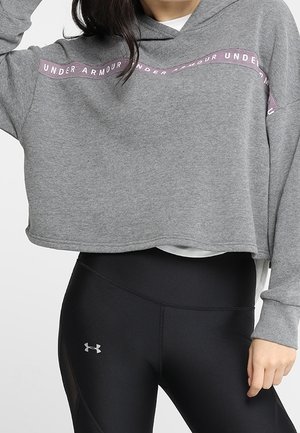 Frau trägt einen grauen, kurz geschnittenen Hoodie mit pinkem Under Armour-Schriftzug und schwarze Under Armour-Leggings mit silbernem Logo am Oberschenkel.