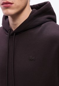 Κοντινό πλάνο ενός ατόμου που φοράει ένα σκούρο καφέ φούτερ με κορδόνια και μικρό λογότυπο κροκοδείλου στο στήθος.