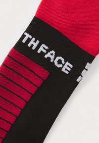 Punane ja must sokk, millel on vertikaalne triibuline disain ning mustal osal on valgest kangast kudumislogoga "THE NORTH FACE".