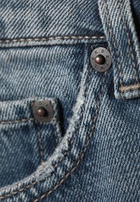 Denim fabric in light blue with contrasting stitching. Features metal buttons with engraved text. Close-up of pocket and seam details.