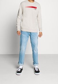 Beige sweatshirt med röd grafisk text på bröstet, parat med ljusblå uppvikta jeans och svarta sneakers med vita sulor och snören.