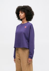 Purple cropped sweatshirt with a round neck, long sleeves, and a small red logo on the chest. Textured fabric paired with beige pants.