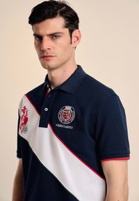Polo marino con un panel blanco diagonal y acentos en rojo y verde, logotipo bordado y mangas cortas. Material de algodón texturizado.