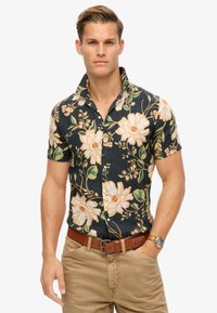 Camicia a maniche corte con bottoni in tessuto scuro e grandi motivi floreali crema e verdi. Abbinata a pantaloni beige e una cintura marrone.