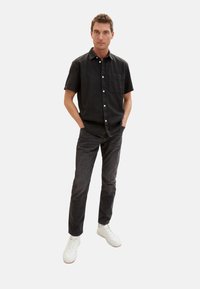 Camicetta nera a maniche corte con bottoni, dotata di taschino sul petto, abbinata a jeans scuri e sneakers bianche. Design semplice e casual con una vestibilità contemporanea.
