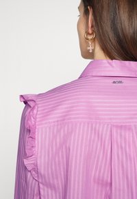 Femme aux cheveux bruns portant un chemisier rose rayé avec un détail à volants sur l’épaule et une boucle d’oreille créole dorée avec des perles roses.