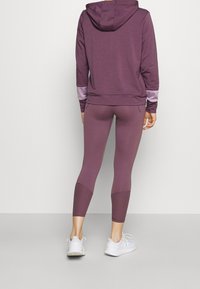 Fialová mikina s kapucí a kontrastními fialovými akcenty na rukávech, v kombinaci s odpovídajícími fialovými legínami. Viditelné bílé sportovní boty.