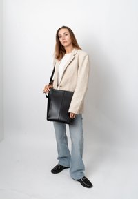 Jeune femme en blazer beige, chemise blanche, jean clair, mocassins noirs, tenant un grand sac à épaule en cuir noir, debout sur fond blanc.