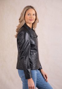 Black leather motorcycle jacket with a wide collar, zippered front, and side pockets. Model wears light blue jeans. Smooth texture.