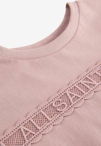 Lyserød bomulds T-shirt med rund halsudskæring, der har en dekorativ blondekant med bølgede kanter og ordet "SAINT" broderet i midten.
