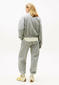 Mujer de pie de espaldas, vistiendo pantalones de chándal grises, sudadera a juego, camiseta con rayas amarillas debajo de la sudadera y zapatillas amarillas y blancas.