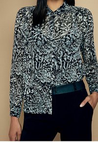 Blusa floreale nera e blu con maniche lunghe, chiusura a bottone e una texture morbida e trasparente. Abbinata a una fascia scura in vita.