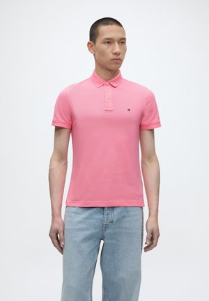 Homme debout portant un polo rose à manches courtes et un jean bleu clair sur un fond blanc uni.