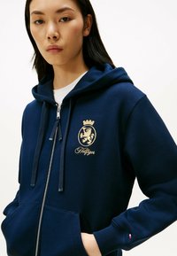 Marineblå lynlås hættetrøje lavet af blødt stof, med et guld løveemblem og "Hilfiger" tekst. Inkluderer snore og frontlommer.