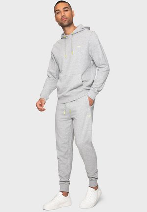 Grauer Hoodie mit Kapuze und Fronttasche, kombiniert mit passenden Jogginghosen. Mit neongelben Kordeln und Logo-Details. Weiße Sneaker.