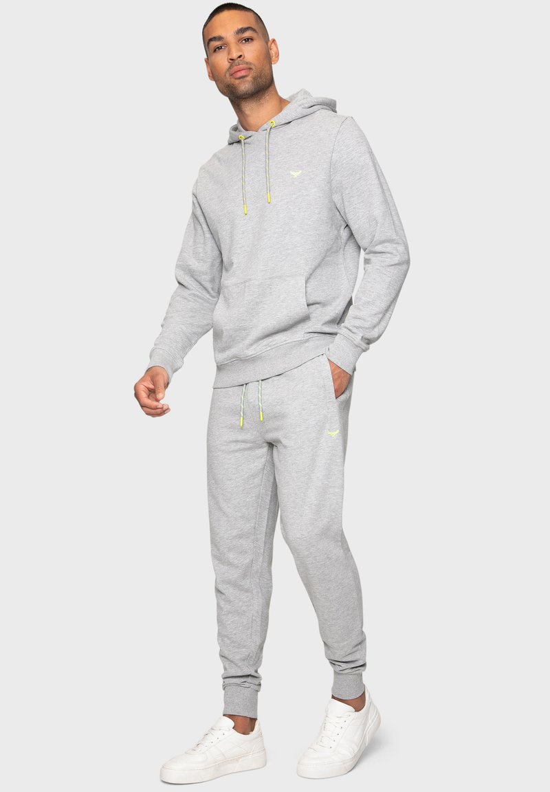 Grijze sweatshirt met een capuchon en een voorzak, gecombineerd met bijpassende joggers. Voorzien van neongele trekkoorden en logo-accenten. Witte sneakers.