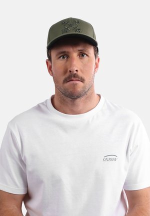 Casquette verte avec un design brodé, portée avec un t-shirt blanc uni comportant un petit logo. Le tissu semble lisse et la coupe est décontractée.