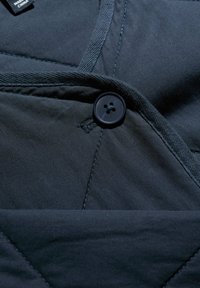 Tumesinine tepitud kangas ümarate kaelusega, millel on must neljarealine nupp ja nähtavad õmbluse detailid.