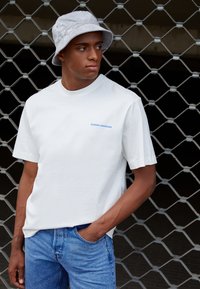 Witte katoenen t-shirt met blauwe tekst "QUEENS BOROUGH", gecombineerd met blauwe spijkerbroek en een lichtgrijze bucket hat. Model poseert tegen een rasterachtergrond.