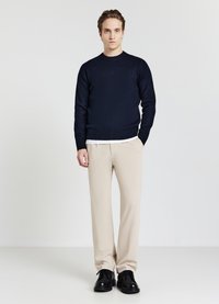 Dunkelblaues Strickpullover mit Rundhalsausschnitt, kombiniert mit beigen Hosen und schwarzen Schuhen. Der Pullover hat gerippte Bündchen und einen gerippten Saum.