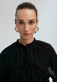 Woman with pulled-back hair wearing large gold hoop earrings and a high-neck black blouse with gathered detailing, facing forward.