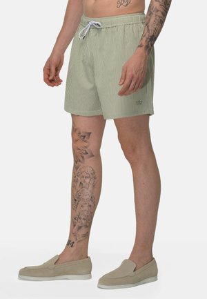 Man draagt beige-wit gestreepte shorts met een wit trekkoord en beige instapschoenen, toont tatoeages op linkerarm en been.
