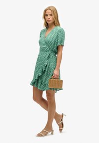 Vestido verde con mangas cortas, patrón floral blanco, dobladillo con volantes y un lazo en la cintura. Combinado con sandalias blancas y una cesta de mimbre.