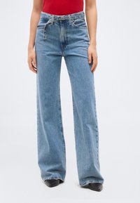 Γυναίκα που φοράει ψηλόμεσα τζιν παντελόνια με φαρδιές μπατζάκια σε ανοιχτό μπλε χρώμα, με ζώνη με τρουκς, κόκκινο τοπ και μαύρα μυτερά παπούτσια, μπροστά σε απλό φόντο.