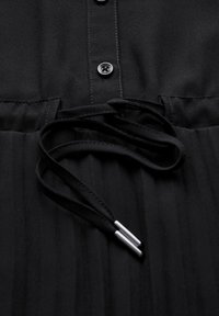 Schwarzes Kleid mit durchgeknöpftem Vorderteil, aus weichem Stoff und mit plissierter Textur. Bänder formen eine taillierte Silhouette und sind mit metallischen Akzenten an den Enden versehen.