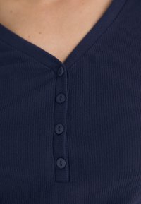 Marineblauw geribbeld top met een V-halslijn en vier knopen aan de voorkant. De stof heeft een fijne textuur met een subtiele rek.