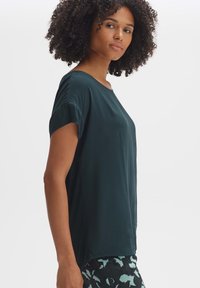 Donkergroene korte mouw top met een ontspannen pasvorm, ronde halslijn en een gladde textuur. Bevat zijsplitsen voor extra comfort en bewegingsvrijheid.