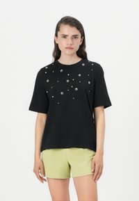 Zwarte T-shirt met korte mouwen en versierde ronde halslijn met verspreide kraalachtige details. Gecombineerd met lichtgroene shorts.
