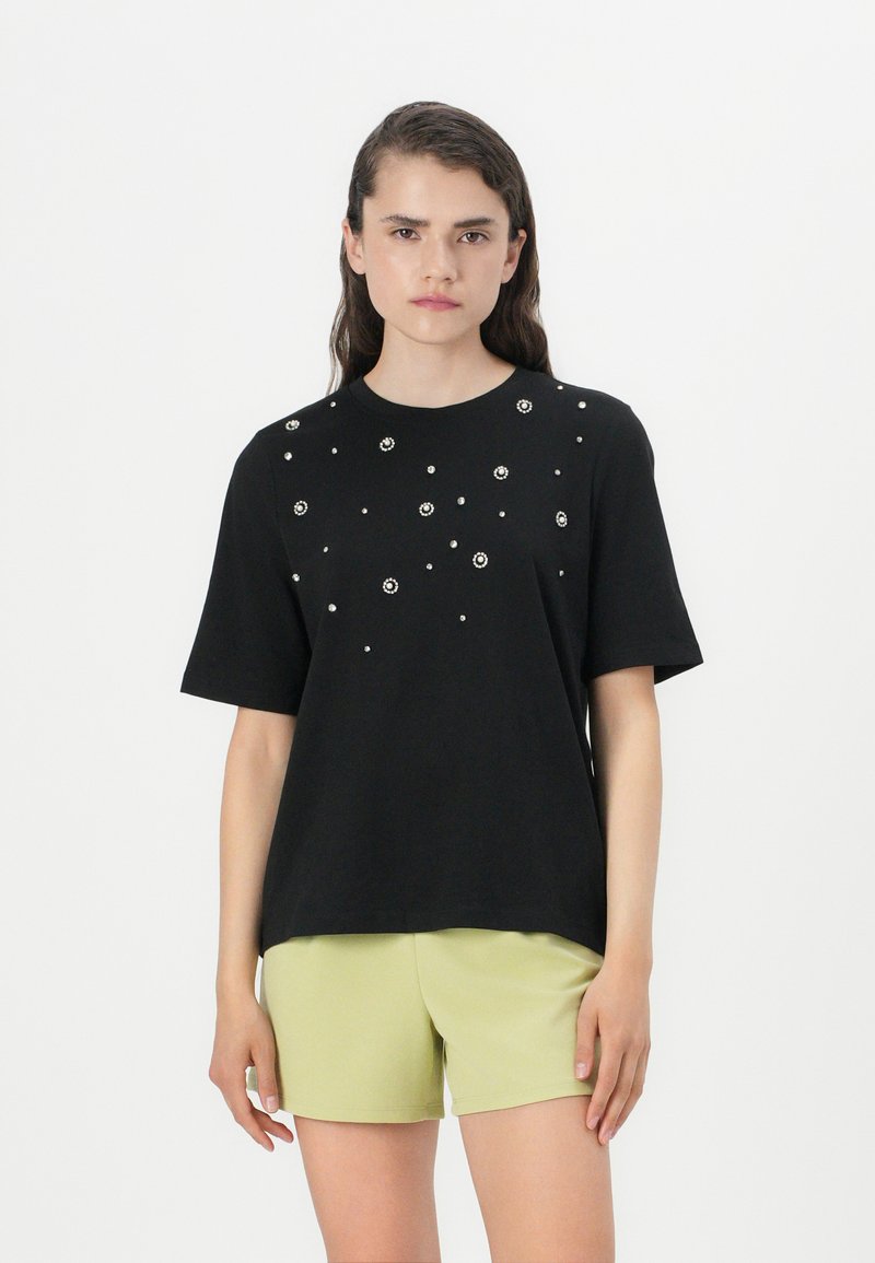 Zwarte T-shirt met korte mouwen en versierde ronde halslijn met verspreide kraalachtige details. Gecombineerd met lichtgroene shorts.