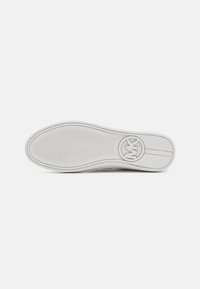 Balta sportbačių pado su tekstūruota sukibimo raštų ir apvaliu logotipu netoli kulno nuotrauka ant neutralios šviesios fono.