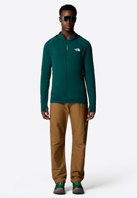 Veste à zip légère verte avec capuche, présentant des accents jaunes, associée à un pantalon marron et des chaussures vertes/grises, modèle portant des lunettes de soleil.